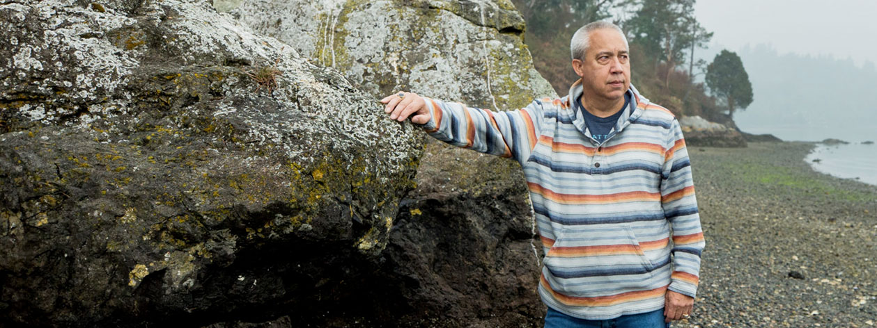 A man stands against a seashore boulder, with his right arm perched onto the boulder, looking out at the water