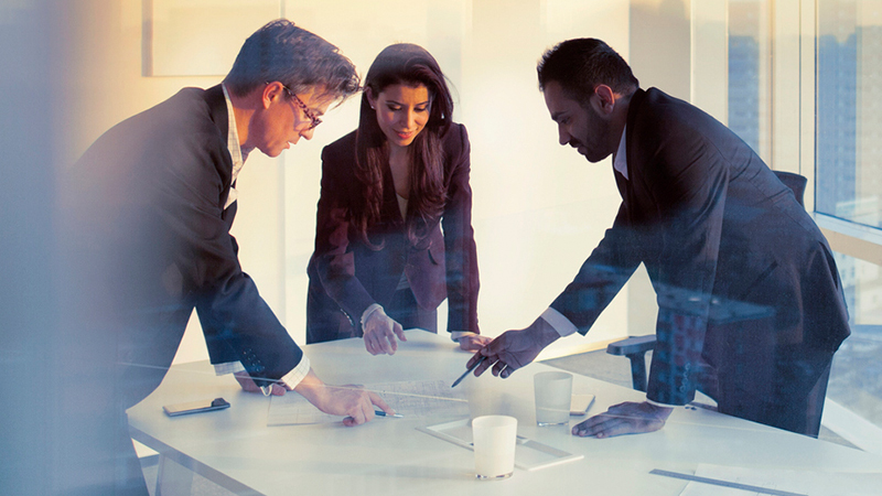 Two business men and a business woman work together, standing over papers on a conference table