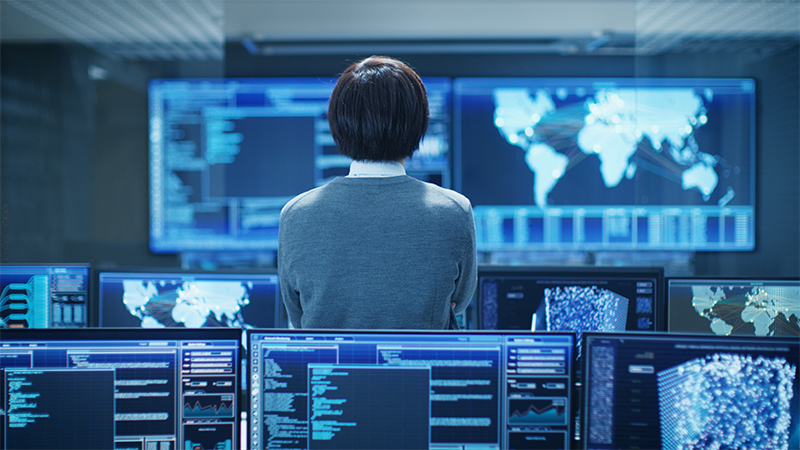 back of woman surrounded by computer screens in dark blue room